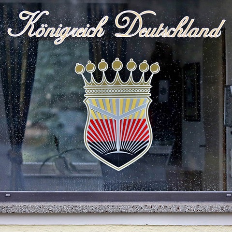 Foto eines Fensters, auf welchem ein Emblem und darüber ein Schriftzug "Königreich Deutschland" in Schreibschrift angebracht ist. Das Emblem zeigt ein Wappen, das eine Krone trägt. Es gliedert sich in schwarz, rot, goldene Flächen (von unten nach oben), wobei sich vom schwarzen "Grund" helle Strahlen nach oben richten.