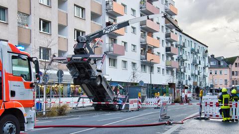 Blick in eine Straßenflucht: Der Arm eines Kranes, der dort steht, ist in Richtung Boden gefallen, während das Bodenteil des Kranes auf einer Seite in die Höhe gekippt ist. Drumherum stehen Feuerwehrmänner und ein Wagen.