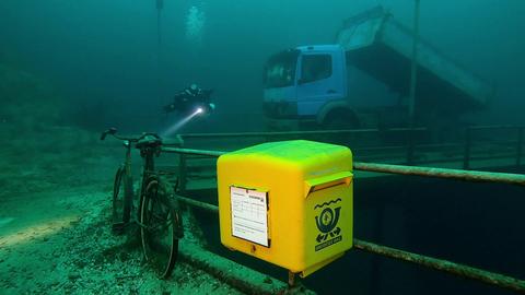 Ein gelber Briefkasten der Marke Eigenbau ist im Kreidesee im Hemmoor (Landkreis Cuxhaven) in einer Tiefe von 19 Metern unter Normalnull angebracht. Daneben steht ein Fahrrad, im Hintergrund ist ein Lkw zu sehen.