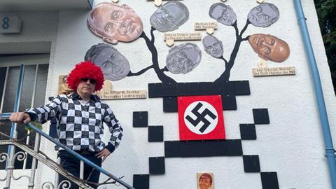Künstler Artur Klose steht auf der Treppe vor seinem Haus neben seinem Kunstwerk, das unter anderem ein Hakenkreuz zeigt