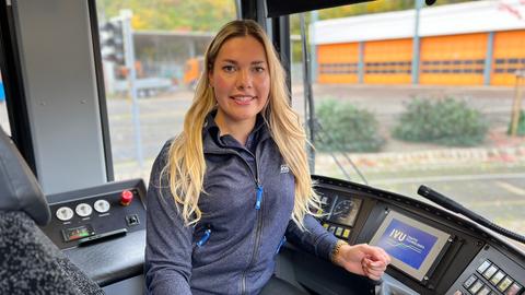 Eine Frau sitzt im Führerhaus einer Straßenbahn und lächelt in die Kamera.