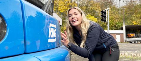 Eine Frau beugt sich zur Vorderseite einer Straßenbahn und streichelt die blau lackierte Oberfläche mit der Signatur "KVG".