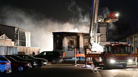 Feuerwehrleute und ein Feuerwehrauto mit ausgefahrener Drehleiter stehen vor der qualmenden Halle.