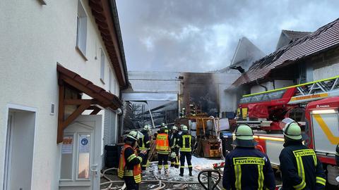 Feuerwehrleute und ein Feuerwehrauto stehen auf dem Hof einer abgebrannten Lagerhalle, auf dem Boden weißer Löschschaum.