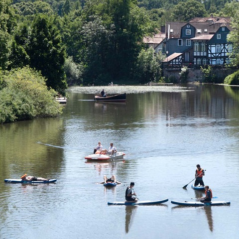 Menschen auf Stand-Up-Paddleing-Boards und in kleinen Booten auf der Lahn. Am grünen Ufer ein Fachwerkhaus.