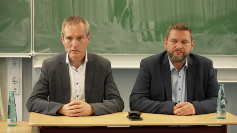 Zwei Männer sitzen an einem Tisch vor einer Schul-Tafel