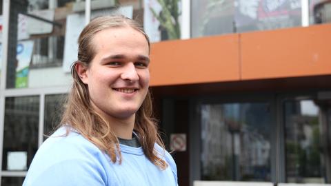 Ein junger Mann mit langen blonden Haaren in einem blauen Pullover steht vor einer Glasfassade.