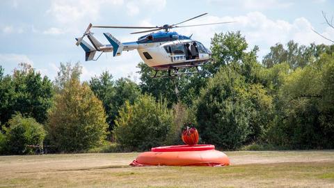 Ein Löschhubschrauber schwebt über einer Wiese und nimmt einen roten Tank ans Seil.