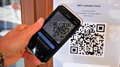 Eine Hand hält ein Smartphone vor einen QR-Code der Luca-App, der an einer Tür angeschlagen wurde.