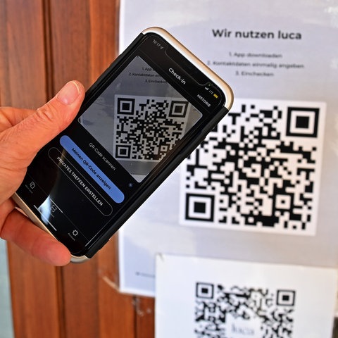 Eine Hand hält ein Smartphone vor einen QR-Code der Luca-App, der an einer Tür angeschlagen wurde.