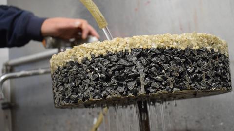 Ein rundes Stück Asphalt aus hellen und dunklen Schichten, oben kommt Wasser aus dem Gartenschlauch darauf, unten sickert das Wasser durch.