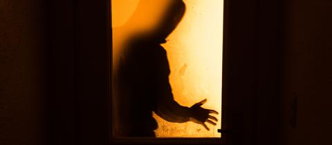 Ein Mann steht bedrohlich hinter einer Tür und ist nur als Schatten sehen. 