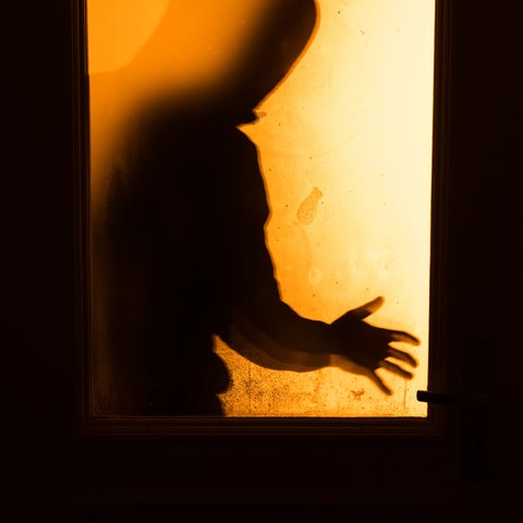 Ein Mann steht bedrohlich hinter einer Tür und ist nur als Schatten sehen. 