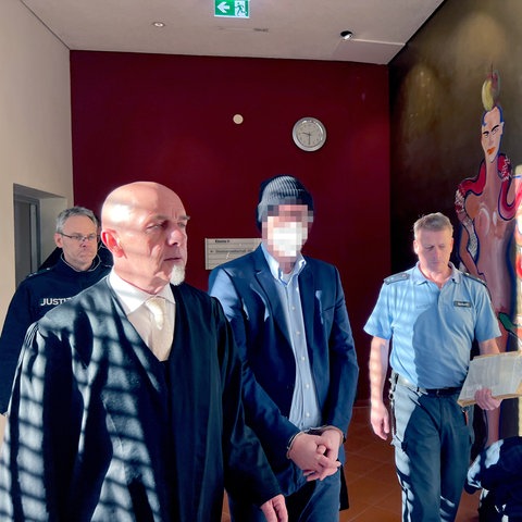 Der Angeklagte (mit Mütze) betritt in Begleitung seines Verteidigers (links) und eines Justizbeamten den Verhandlungssaal im Landgericht Fulda. 