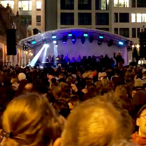 Masse von Menschen vor eine bunt bestrahlten Bühne auf einem städtischen Platz.