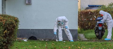 Männer in weißen Schutzanzügen und Handshcuhen suchen den Rasen vor einem Haus nach Spuren ab.