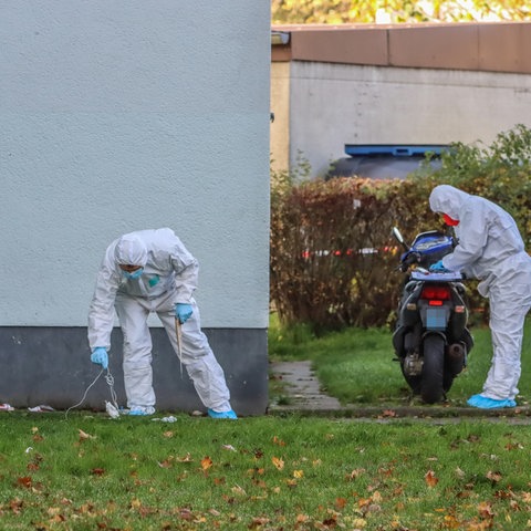 Männer in weißen Schutzanzügen und Handshcuhen suchen den Rasen vor einem Haus nach Spuren ab.