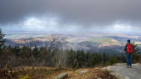 Ein Wanderer, ausgestattet mit einem leuchtend roten Rucksack, steht auf dem Gipfel des Feldbergs im Taunus und blickt in die Ferne.