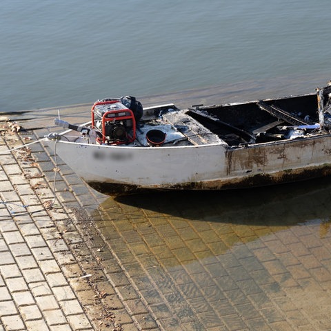 Ein ausgebrannte Motorboot am Ufer einer Wasserfläche.