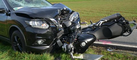 Ein schwarzes Motorrad, das unter einem schwarzen Pkw geraten ist - beide Fahrzeuge sind teils schwer beschädigt.