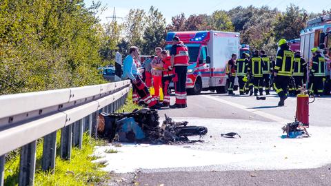 Im Vordergrund liegt auf dem Asphalt der Landstraße das ausgebrannte Gerippe eines Motorrads. Im Hintergrund lehnen Ersthelfer an der Leitplanke. Dahinter stehen die Rettungswagen. 