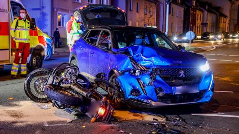 Das beschädigte Motorrad liegt neben dem Unfallauto, dahinter Rettungskräfte.