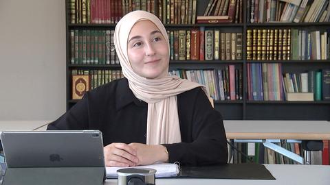 Muslima mit Kopftusch sitzt am Schreibtisch, im Hintergrund sieht man ein Bücherregal.