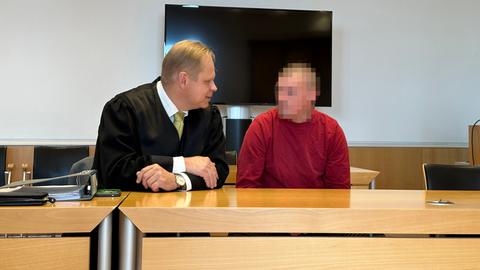 Ein Mann in rotem Pullover sitzt in einem Gerichtssaal, links neben ihm sein Anwalt in schwarzer Robe.