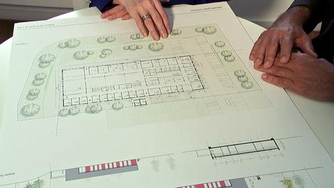 Eine Mann und eine Frau sitzen an einem Tisch. Vor ihnen liegt ein Entwurfsplan eine Gebäudes.