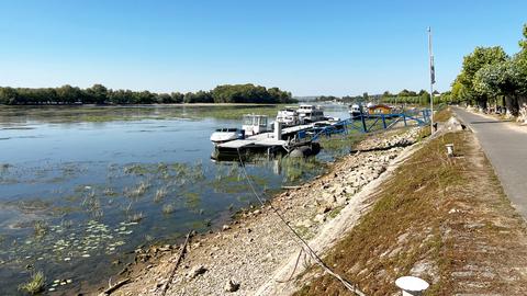 Flussufer mit einem Anleger und einigen kleineren Schiffen. Das Flusswasser steht so niedrig, dass die ganze Böschung des Flussufers trocken liegt und sichtbar ist.