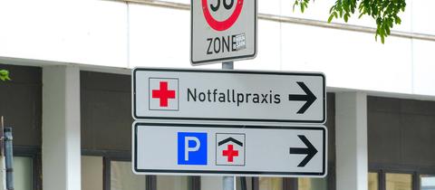 Schilder, die vor einem Krankenhaus stehen. Sie zeigen den Begriff "Notfallpraxis" und zwei Icons für Krankenhaus und Parkplatz