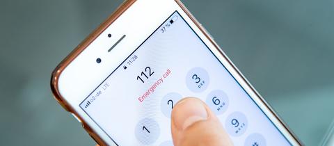 Die Notrufnummer 112 auf dem Screen eines Smartphones. Daneben ein Daumen, der auf dem Screen liegt.