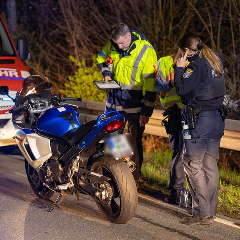 Eine Polizistin und ein Gutachter stehen mit einem Notizblock neben einem Motorrad, im Hintergrund ein Feuerwehrauto. 