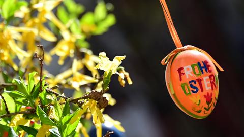 Ein orangefarbenes Osterei mit dem Schriftzug "Frohe Ostern" hängt an einem Forsythienzweig.