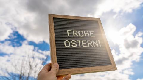 Im Vordergrund hält eine Hand eine Tafel hoch, auf der frohe Ostern steht. Im Hintergrund ein wolkiger Himmel.