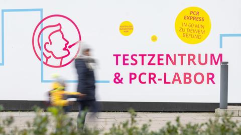 Zwei Menschen (unscharf laufen an einer Plakatwand vorbei, auf welcher für PCR-Tests mit Ergebnis innerhalb von 60 Minuten geworben wird.