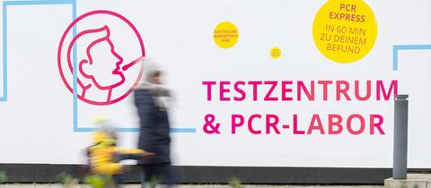 Zwei Menschen (unscharf) laufen an einer Plakatwand vorbei, auf welcher für PCR-Tests mit Ergebnis innerhalb von 60 Minuten geworben wird.