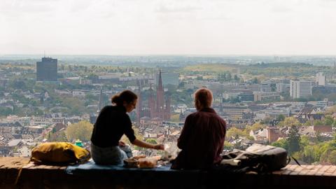 Zwei Personen sitzen beim Picknick auf einer Mauer. Bei schönem Wetter hat man einen weiten Blick über Wiesbaden.