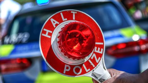 Im Bildvordergrund eine Kelle mit der Aufschrift "Halt Polizei". Im Hintergrund - unscharf - ein Polizeiwagen.