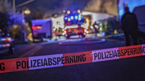 Absperrband mit "Polizeiabsperrung" im Vordergrund des Bildes. Im Hintergund, unscharf, Feuerwehr im Einsatz vor einem Gebäude.