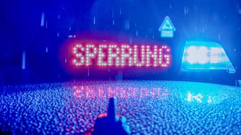 Im Bildvordergrund ein Blaulicht mit Leuchtanzeige "Sperrung" auf einem Autodach. In Hintergrund - unscharf - eine Straße im Regen.