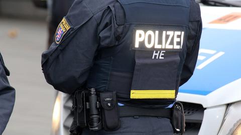 Das Foto zeigt einen Polizisten von hinten im Bildausschnitt wie er neben einem Polizeiauto steht. Deutlich ist die Aufschrift "Polizei" auf seinem Rücken zu sehen.