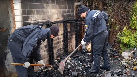 Zwei Polizisten stehen in einer erloschenen Brandstelle und schauen mit Schaufeln nach Spuren.
