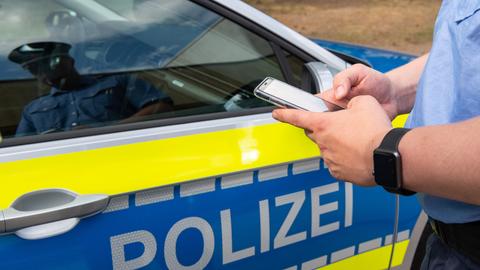Polizist tippt etwas in ein Smartphone, im Hintergrund steht ein Streifenwagen
