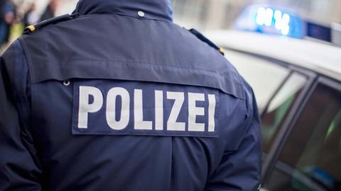 Polizist von hinten, auf der Jacke steht in großen Lettern "Polizei". Daneben ein Polizei-Auto mit Blaulicht. Sujetbild.