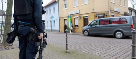 Bildvordergrund (scharf): Polizist mit Maschinengewehr steht an einer Straße. Bildhintergrund (leicht unscharf): Gebäude mit Eckkneipe, davor steht ein Leichenwagen.