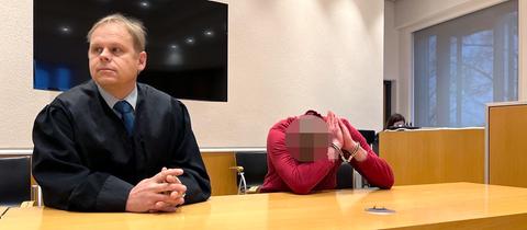 Zwei Männer sitzen am Tisch. Links ein Mann in Anwaltsrobe, rechts ein Mann in rotem Pulli, der sein Gesicht in der Armbeuge versteckt und Handschellen trägt.