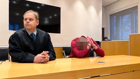Zwei Männer sitzen am Tisch. Links ein Mann in Anwaltsrobe, rechts ein Mann in rotem Pulli, der sein Gesicht in der Armbeuge versteckt und Handschellen trägt.