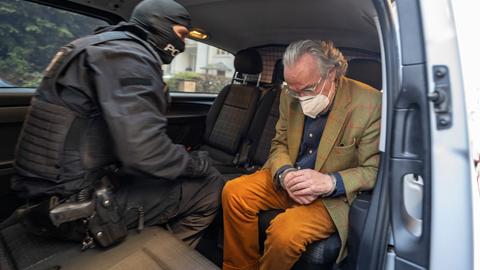 Heinrich XIII. Prinz Reuß in einem Polizeiauto mit Handschellen neben einem Polizisten.