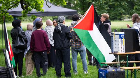 Das Bild zeigt Teilnehmende eines pro-palästinensischen Protestcamps auf dem Campus der Goethe Universität. Sie stehen unter einem auf einer Wiese aufgebauten Pavillon, tragen Palästinensertücher und schwenken die palästinensische Fahne.